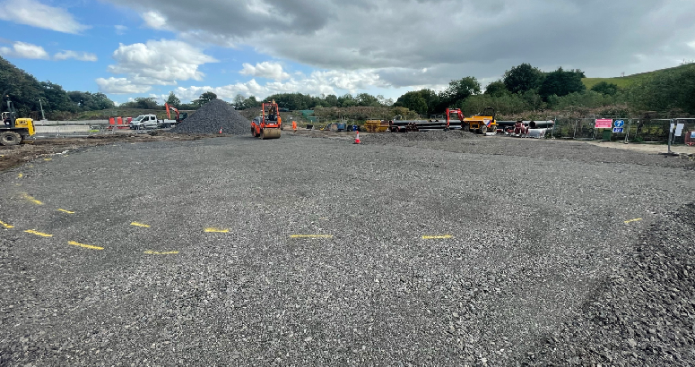 Image showing a large construction site with various heavy machinery including excavators, working on gravel-covered land under a cloudy sky.