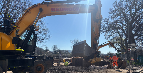 A digger sits at the forefront with construction workers in the background and a blue sky overhead.