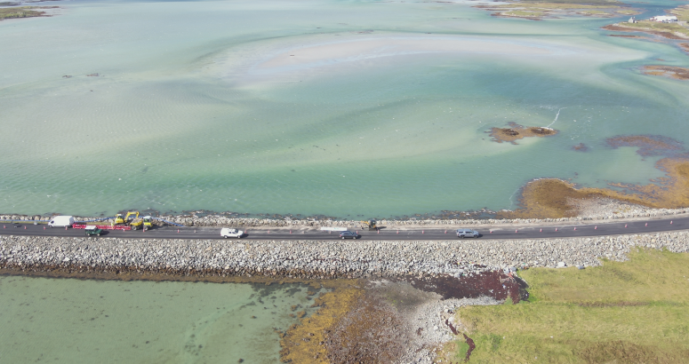 Aerial view of a coastal road with cars and contractor vehicles near a large, shallow turquoise and green bay with visible aquatic plants and sand patches.