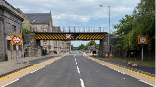 View from the middle of Tay Street road. Buildings line the pathway on the left and trees on the right. A railway bridge can be seen crossing over the road.