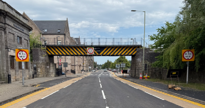 View from the middle of Tay Street road. Buildings line the pathway on the left and trees on the right. A railway bridge can be seen crossing over the road.