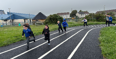Children running along a running track. Grass surrounds the track and buildings can be seen in the background. The sky is grey.