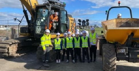 Children stand in high viz and hard hats in front of a digger and dumper. The sky is blue.