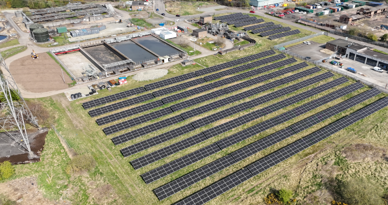 An aerial view of a solar farm with panels mounted on top of grass. In the background of the image is a waste water treatment works.