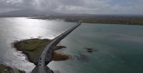 Aerial view of a coastal road crossing over a narrow strip of land with shimmering blue waters on either side, leading towards distant mountains under a cloudy sky.