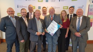 Group of nine people posing with an award at an event. Some are smiling and holding a certificate and a trophy. The setting suggests a professional or corporate event.