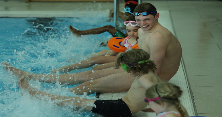 Duncan smiling while sitting pool side with children. All are kicking the water to make a splash.