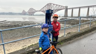 Two young children pose beside bright blue water refill fountain with several large bridges in the background