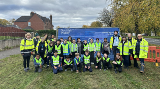 Group of people of various ages wearing safety vests, standing in front of a Scottish Water truck, in a suburban area.