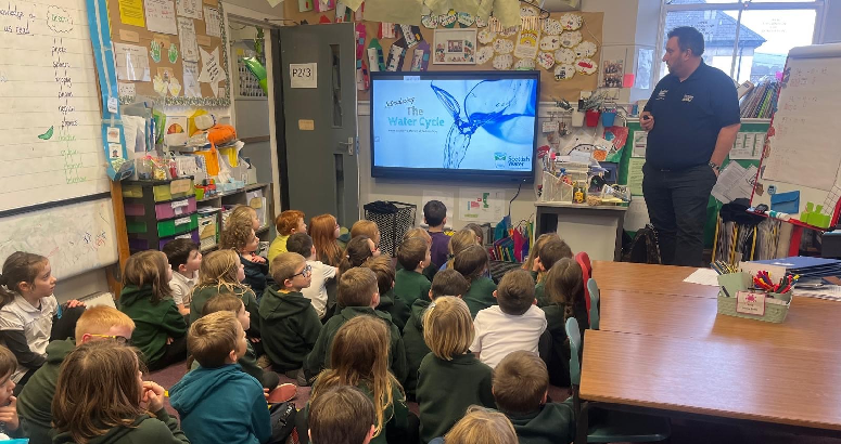 A group of young students in green uniforms sit on the floor of a colourful classroom, watching a presentation on a screen at the front. An adult stands nearby, delivering the talk. The room is filled with educational materials, posters, and decorations.